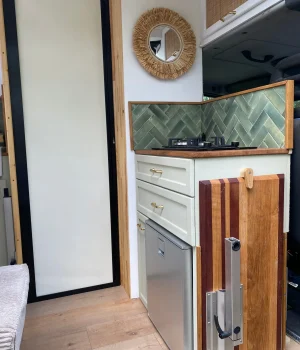 Vue intérieure depuis la porte latérale avec cuisine équipée, réfrigérateur, table escamotée et meubles hauts en cannage.